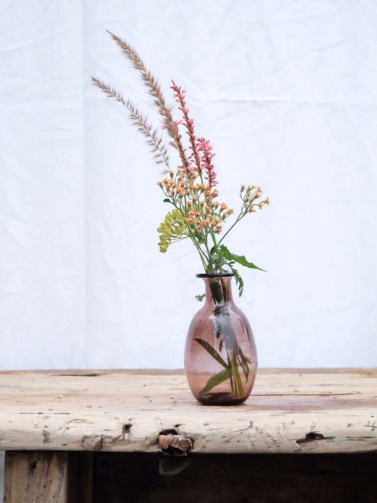 Small raspberry coloured vase with spring flowers sitting on a vintage wooden table. Handblown in Paris by La Soufflerie.
