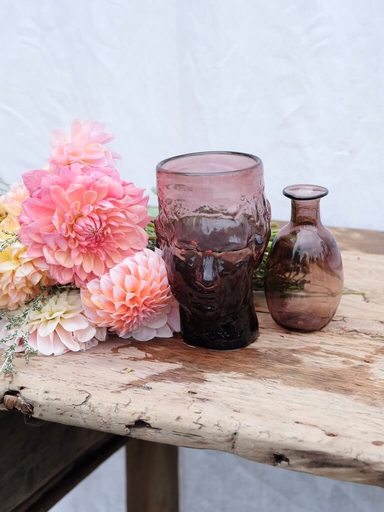 Flowers and two raspberry coloured vases sitting on a vintage wooden table. One is in the shape of a womans head and the other is a small lipped vase. Both are handblown in Paris by La Soufflerie.
