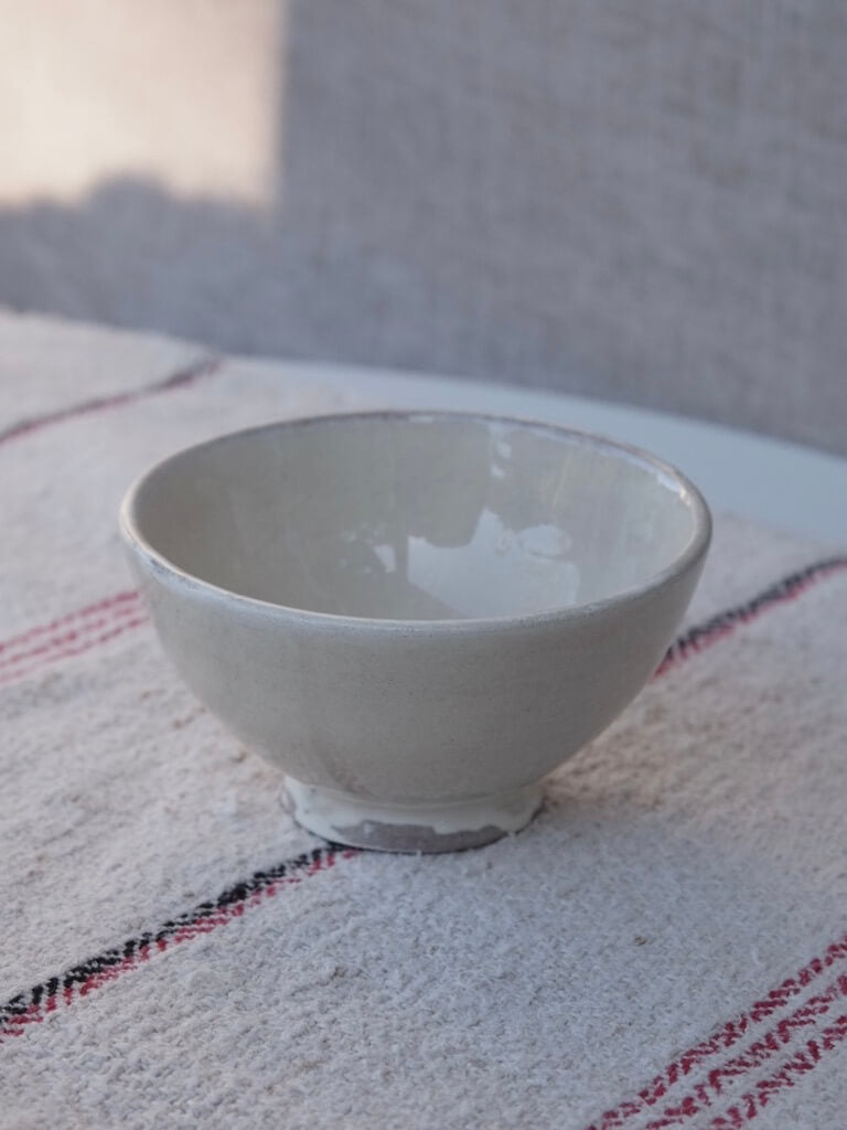 small footed cream coloured bowl with an uneven glaze. It is photographed on a table with a vintage hungarian table runner with black and red stripes