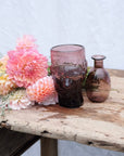 Flowers and two raspberry coloured vases sitting on a vintage wooden table. One is in the shape of a womans head and the other is a small lipped vase. Both are handblown in Paris by La Soufflerie.